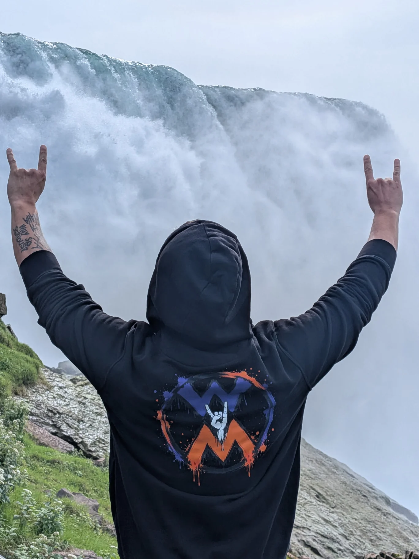 Person raising both arms in front of a waterfall, wearing a black hoodie with the purple and orange Worth More Movement logo
